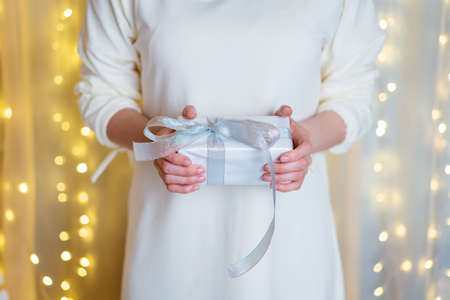 Christmas gift in human hands holiday wallpaper poster conceptual painting with white wall background and lights garlands selective focusの写真素材