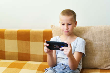 Close-up of cute little boy using smartphone looking at screen, curious child holding phone in hands, sitting on sofa at home alone, playing game on mobile device, watching cartoons online, chattingの写真素材