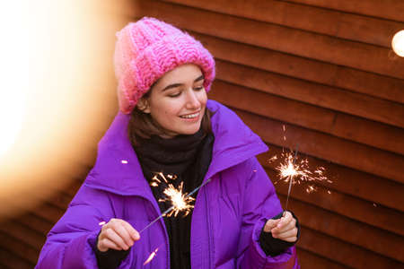 Happy young woman in outerwear with sparklers on the street on the background of a wooden wall...