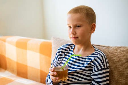 Young calm boy in casual clothes holds fresh juice drink in glass made of glass with a plastic straw, sits on the sofa at home, in an apartment indoors, resting on weekends. People hall concept.の写真素材