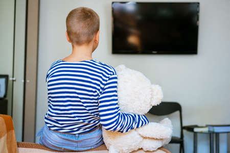 Cute boy and his plush toy bear are watching tv while sitting on the couch in the living room at home. View from the back. Young guy in striped t-shirt and blue jeansの写真素材