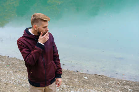 Young bearded man stood alone by lake and looked aside thoughtfully smoking cigarette. Fog over the...