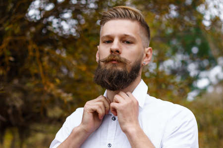 Autumn mood. Pensive young man in white shirt, meditates and walks in park against the backdrop of beautiful autumn trees. Autumn is coming. Autumn story. Yellow leaves in the park. Portrait of a manの写真素材