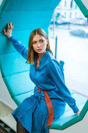 Caucasian woman sitting by window on bench in shape of circle in blue dress on the background of the city. Business woman thinkingの写真素材