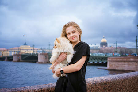 Cheerful and happy girl teenager holds and hugs pomeranian spitz on embankment in city on cloudy day, selective focus. Caucasian young woman with her beloved pet on the river bank in the wind smilingの写真素材