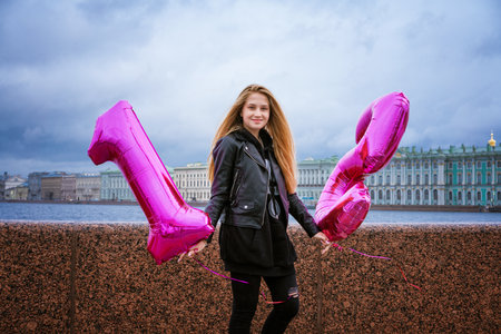 Happy girl holding balloons on the embankment of the city in a leather jacket. The teenager celebrates his birthday in hand with balloons in the form of numbers. On an autumn cloudy dayの写真素材