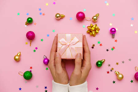 Woman hands holding gift, decorated with ribbon on pink background with christmas balls and multicolored candy stars, copy space. Flat lay, hands and gift box, top view. Christmas concept.の写真素材