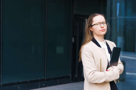 A successful business woman of Caucasian ethnicity with a folder of papers in her hand, in a coat and suit, in glasses stands near a business center and smilesの写真素材