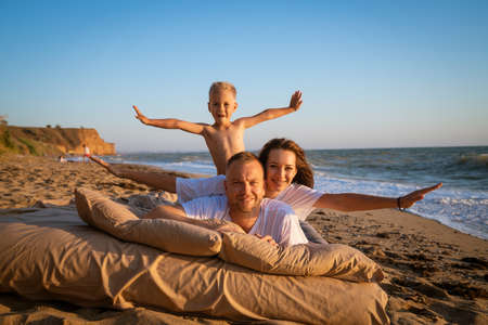 a happy family of three people is resting on a sandy beach. Mom is a brunette with long hair, father and son are blonde with short haircuts. people have fun and pose on the bedの写真素材