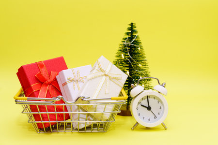 Gift boxes in shopping basket with white alarm clock and small artificial christmas tree on a yellow...
