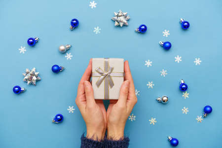 Womans hands holding gift box present, tied with ribbon bow on blue background around christmas tree decor with balls and snowflakes. Christmas, new year, holidays, birthday conceptの写真素材