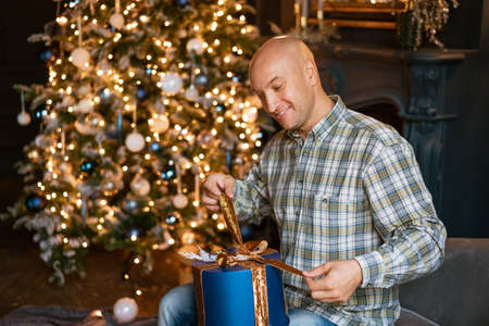 Happy bald man in a shirt opens a Christmas present while sitting on the sofa against the background of a Christmas treeの写真素材