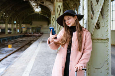 Happy young woman on platform of railway station in pink coat and black hat stands waiting for train and communicates via video communication with relatives online or takes a selfie on a smartphoneの写真素材