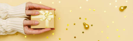 Female hands in pullover, holding christmas gift box decorated with confetti gold stars and christmas balls on a yellow background. Christmas and new year banner.の写真素材