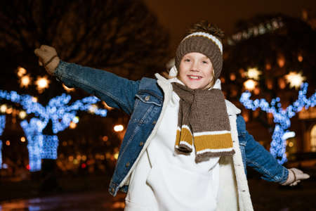 Portrait of joyful caucasian teenager boy standing on street in evening in city lights. Cheerful little male teenager in the city smiling. Snow in the open air. Christmas holidays conceptの写真素材