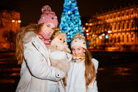Two young girls are smiling and having fun holding spitz dog looking out christmas street decor. Beautiful sisters, decorated christmas tree and lights on background. A magical evening on the streetの写真素材