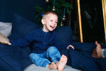 Cute little blond toddler boy in blue sweater is having fun and playing on couch laughing...
