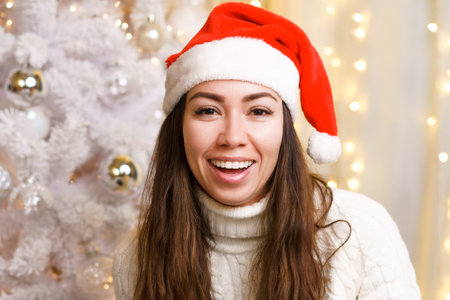 Happy young woman in hat of santa claus in white knitted suite on background of christmas tree with toys and a garland cute smiling looking at the camera. Caucasian cheerful girl in a festive moodの写真素材