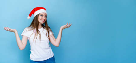 Pretty young woman in santa hat and white t-shirt posing cheerfully on blue background, copy space. Caucasian cheerful girl dancing for joyの写真素材