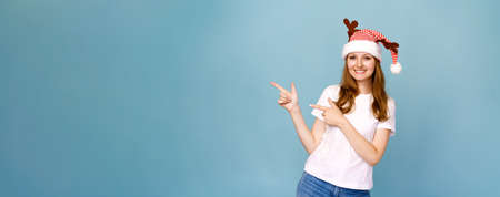 Cheerful santa girl in funny decorative deer antlers, pointing with forefinger to side, smiling with...