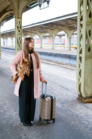 Woman traveler tourist walks with luggage and dog at train station in pink coat. The concept of an...
