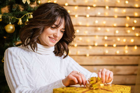 Smiling happy young woman in winter sweater holding golden present while sitting on floor at home against the background of a christmas tree in a cozy living room with a festive interior.の写真素材
