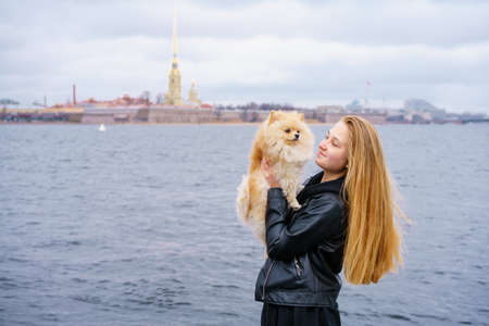 Lifestyle image of happy attractive young woman in black leather jacket walking on the river promenade with her cute spitz dog stroking her head and looking at the camera with a joyful smileの写真素材