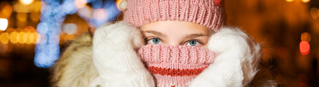 Close-up portrait young girl in knitted hat holds her hands in mittens near her face, covers her face with scarf from cold outside. Girl in evening on christmas street in light of lights and garlandsの写真素材