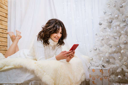 Holidays and technology concept happy smiling girl with smartphone lying in bed at home against background of christmas tree in white knitted sweater. Caucasian young woman enjoying holidays at homeの写真素材