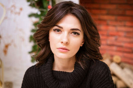 Close-up portrait of woman looking at camera against background of fireplace with christmas decor....