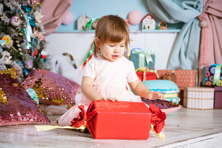 A little girl sits on floor with gifts next to christmas tree in pink dress. Happy caucasian child is enjoying christmas and new year.の写真素材