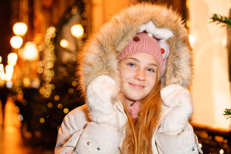 Street portrait smiling beautiful girl on festive christmas street in evening against background of christmas lights hands in mittens. Girl in stylish winter warm clothes. The model looks into camera.の写真素材
