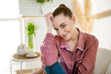 Calm young woman sits on comfortable chair and enjoys calm peaceful relaxation in living room while holding her head. Lazy serene lady meditates, breathes fresh air, relaxes with smile on her face.の写真素材