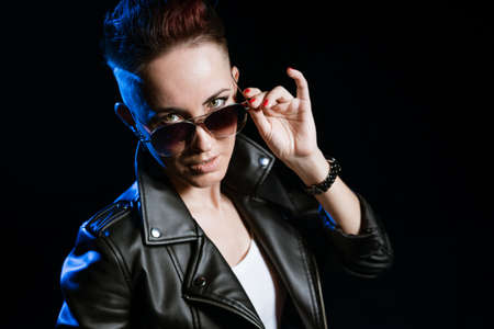 Stylish short haired brunette woman in black leather jacket wearing sunglasses makes stop gesture with hand on black background with blue light. Confused and negative concept.の写真素材
