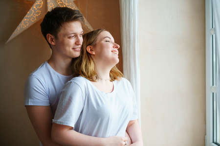 Shot of happy young couple standing together by window while looking away. Handsome man hugs beautiful smiling woman. In a light t-shirt. Romantic time together in the morning at homeの写真素材