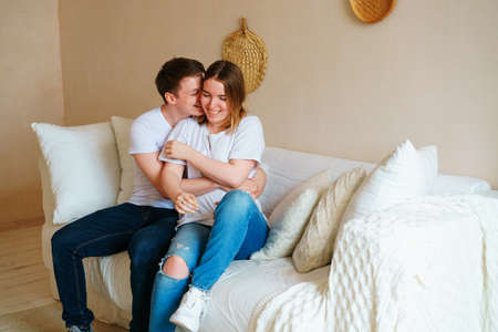 Young couple at home in casual clothes. Guy and girl hug and kiss, having fun, enjoy spending time together, sitting on the couch at home.の写真素材