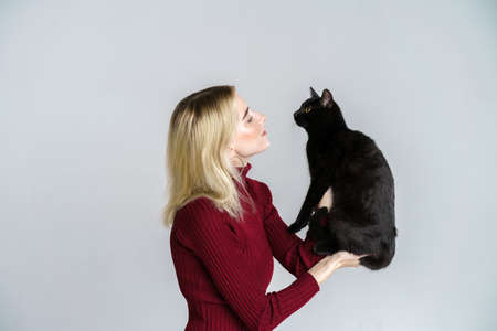 Young caucasian pretty cute blonde in red holding black cat, like baby, isolated on light background background. The girl is babysitting the kitten. Friendship between pet and ownerの写真素材