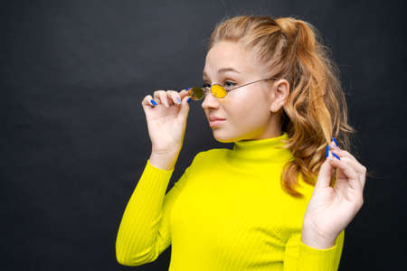 Close-up portrait happy young girl with blond hair, looks through yellow glasses, in a yellow...