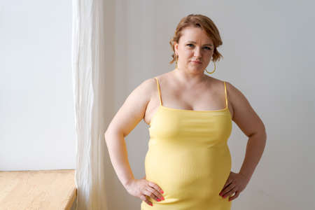 Portrait cheerful attractive young plump plus size woman in yellow tight dress looking at the camera posing indoors by the window with copy space for your informationの写真素材