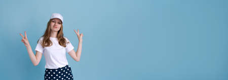 Photo an attractive positively surprised young woman pointing her finger at copy space presenting an advertisement. Caucasian girl shows v-sign on blue background in white t-shirt and cap with mock-upの写真素材