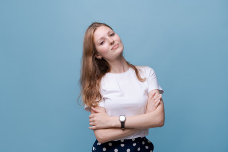 Smiling beautiful millennial girl in white t-shirt stand with arms crossed isolated on blue studio background, happy young woman in looking at camera, posing or castingの写真素材
