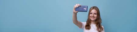 Young female vlogger with surprised face takes picture herself on phone while broadcasting on a blue...