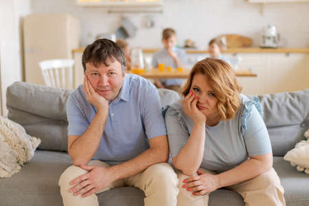 Tired parents suffer from headaches, sit on couch, while their daughter and two sons run in the kitchen and scream, dad and mom feel exhausted by noisy children. Disobedient children conceptの写真素材