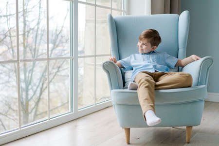 Happy boy in blue shirt relaxes with good thoughts. Peaceful guy is resting in chair at home by window, thinking and dreaming, looking out window. Comfort in apartment, home conceptの写真素材