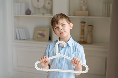 Little caucasian boy is holding hanger in room against background wall with shelves and looks into camera and smiles. The concept of buying, selling and childrens fashion clothing.の写真素材
