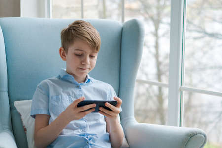 Happy pause. Positive guy with smartphone, listening to music at home or playing games, watching a video online, sitting in a blue chair by the window. Millennial boy enjoying free time at homeの写真素材