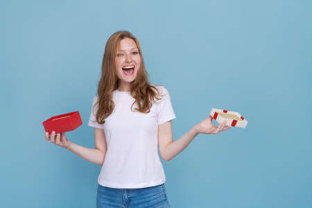 Surprised young caucasian woman holding red gift box and shocked with open mouth standing on blue...