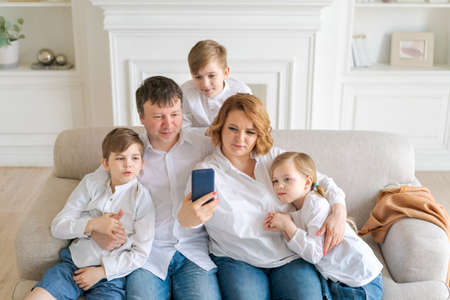 Happy cheerful caucasian family dad mom and kids having fun and using digital phone video call on couch at home. Self-isolation sitting at home social distancing quarantine for coronavirus prevention.の写真素材