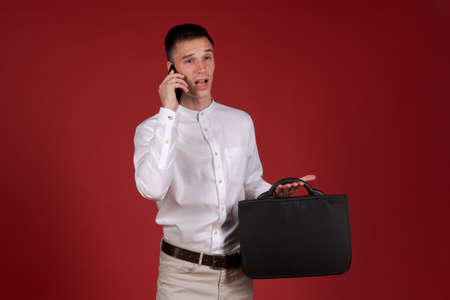 An angry man in white shirt and light trousers with document bag in his hand screams on a mobile phone on the speakerphone. Stressed and depressed guy on a red background. Emotional manの写真素材