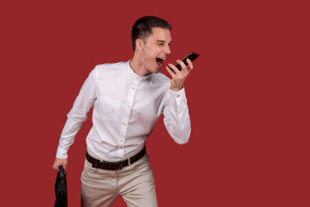 Image young business emotional excited screaming man in white shirt isolated over red background wall screaming into mobile phone handset on speakerphone, emotional workerの写真素材